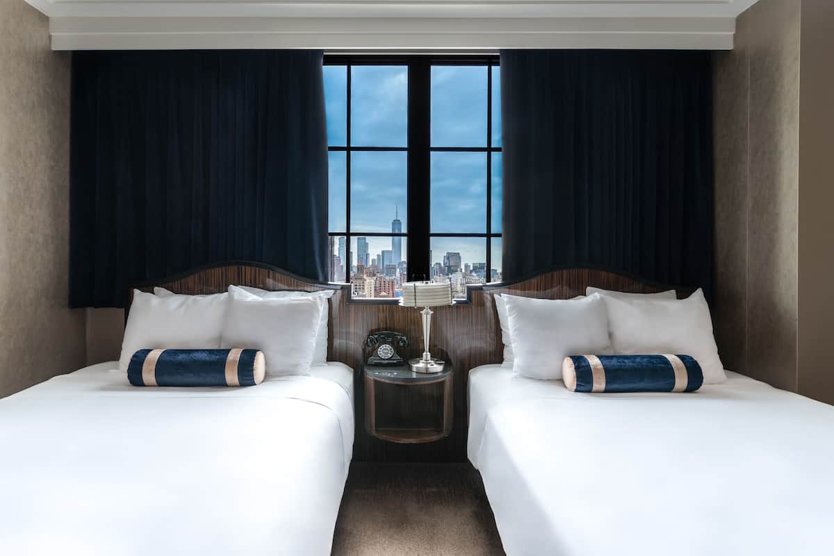 A hotel room with two beds, a nightstand with a lamp, and a view of a city skyline through large windows.