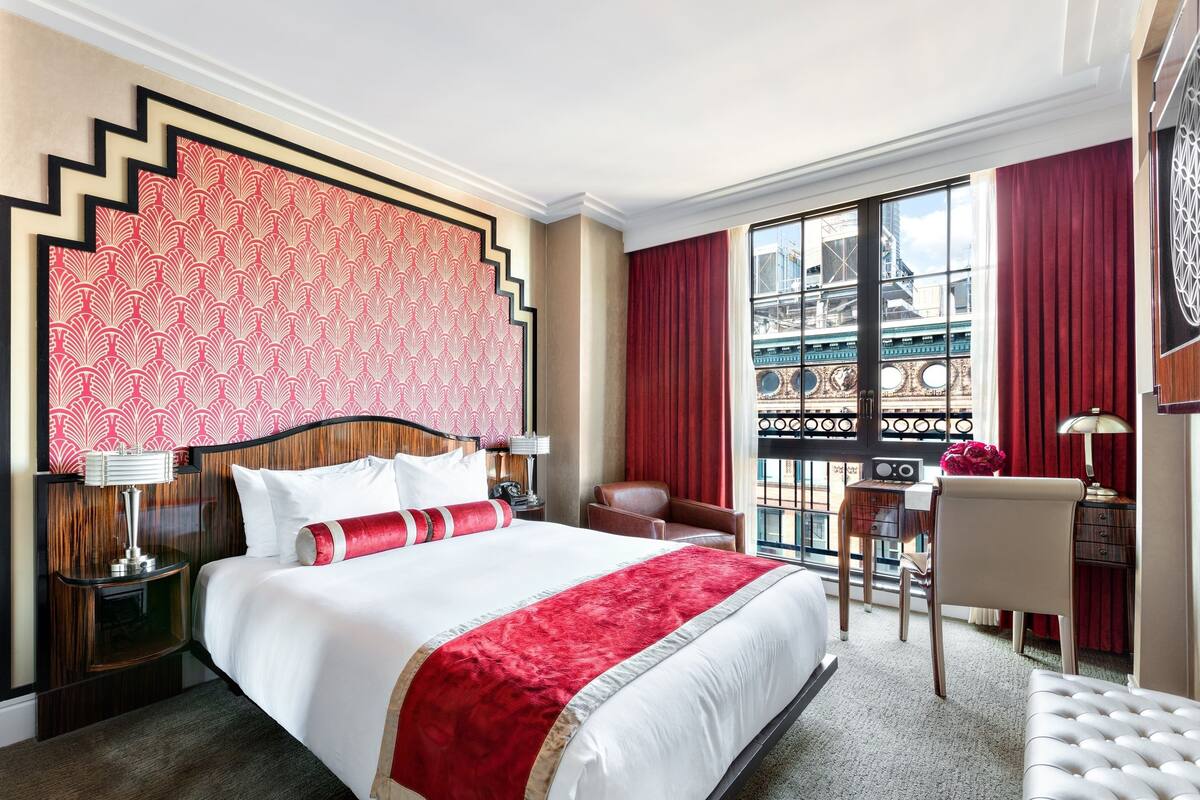 A hotel room with a large bed, a desk, and a chair. There is a window with a view of buildings.