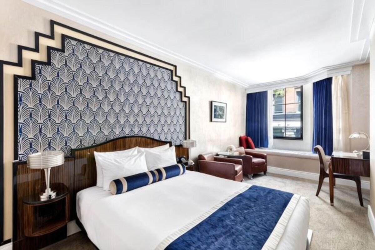 A hotel room with a large bed, a desk, a chair, a nightstand, and a window with curtains.
