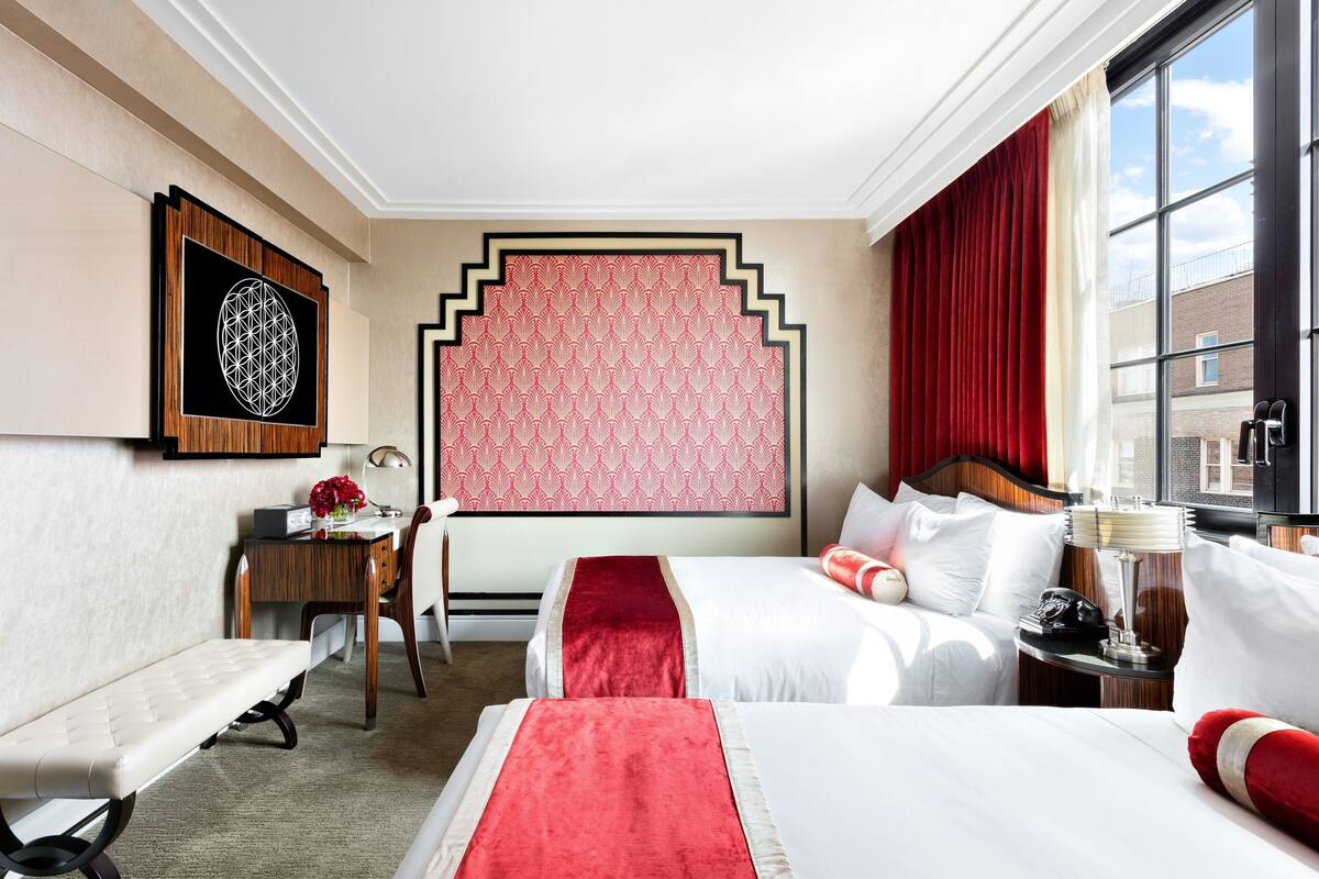 A hotel room with two beds, a desk, a chair, a bench, a large window, and a patterned wall.