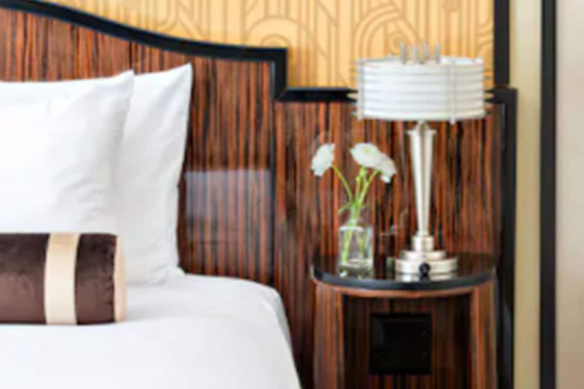 A hotel room with a patterned wallpaper, a wooden headboard, a bed with white bedding, a nightstand with a lamp, and a vase with flowers.