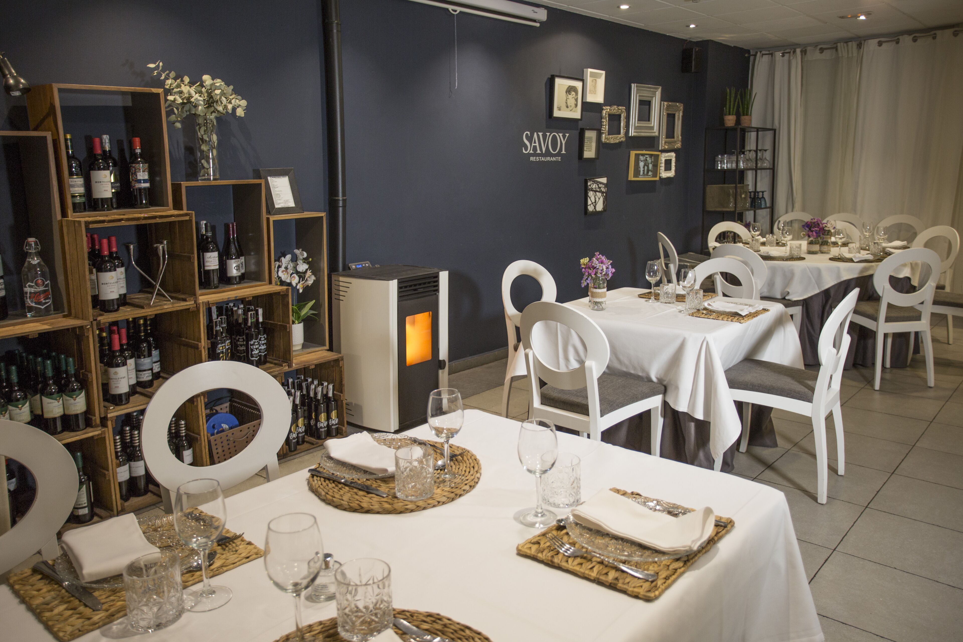 Interior de restaurante com mesas preparadas para refeições, garrafas de vinho em estantes e quadros nas paredes.