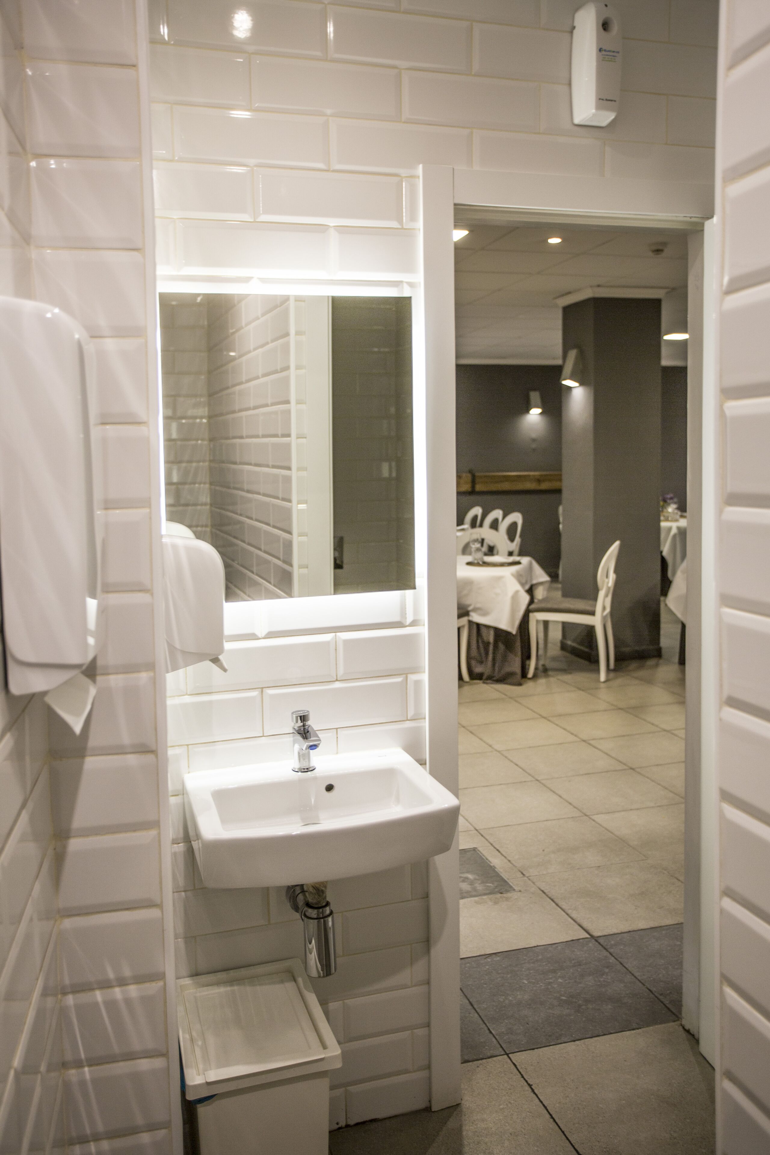 A bathroom with a white sink, mirror, and tiled walls. There is a small trash bin under the sink and a towel hanging on the wall.