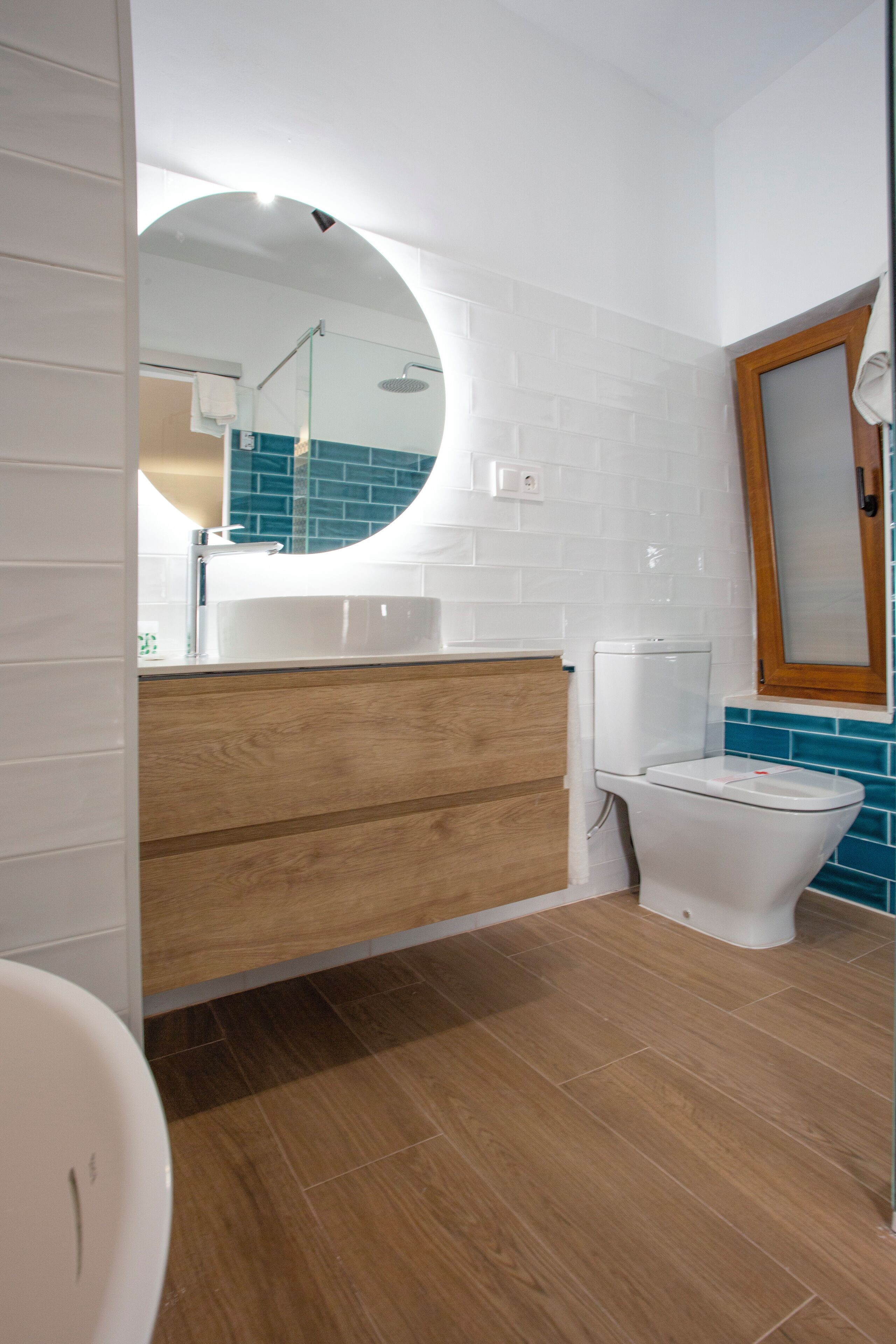 Un baño moderno con tocador de madera, lavabo blanco y espejo redondo.