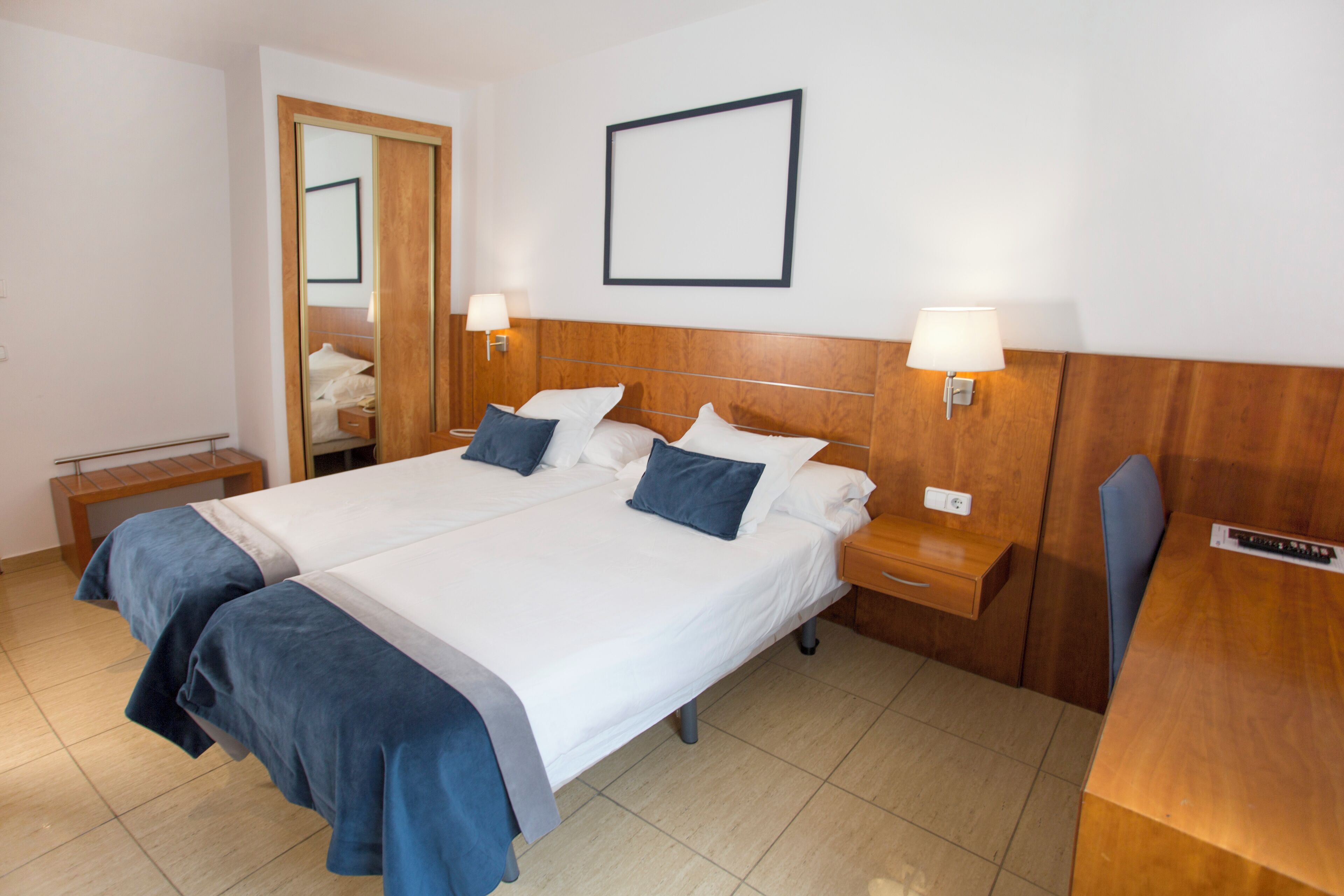 A double bed with a wooden headboard, two bedside tables with lamps, a wooden bench, and a mirror.