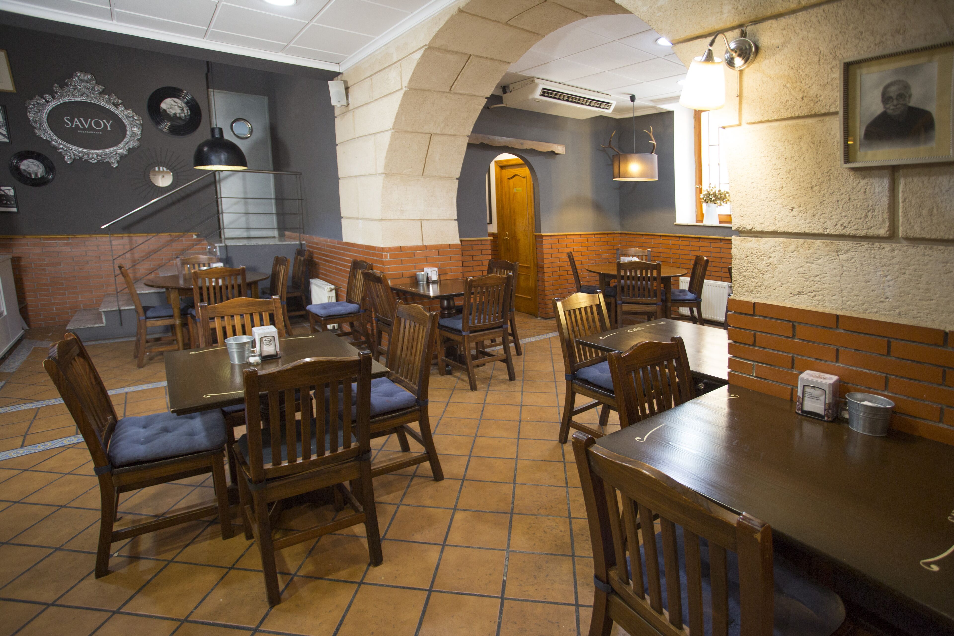 Interior de restaurante con paredes de ladrillo, mesas y sillas de madera, y una escalera que lleva a un nivel superior.