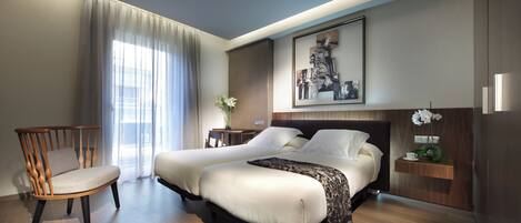 A modern hotel room with a large bed, a wooden chair, a small table with a vase of flowers, and a framed picture on the wall.