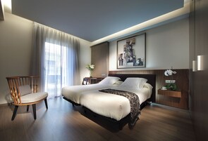 Una habitación de hotel moderna con una cama grande, una silla de madera, una mesita con un jarrón de flores y un cuadro en la pared.