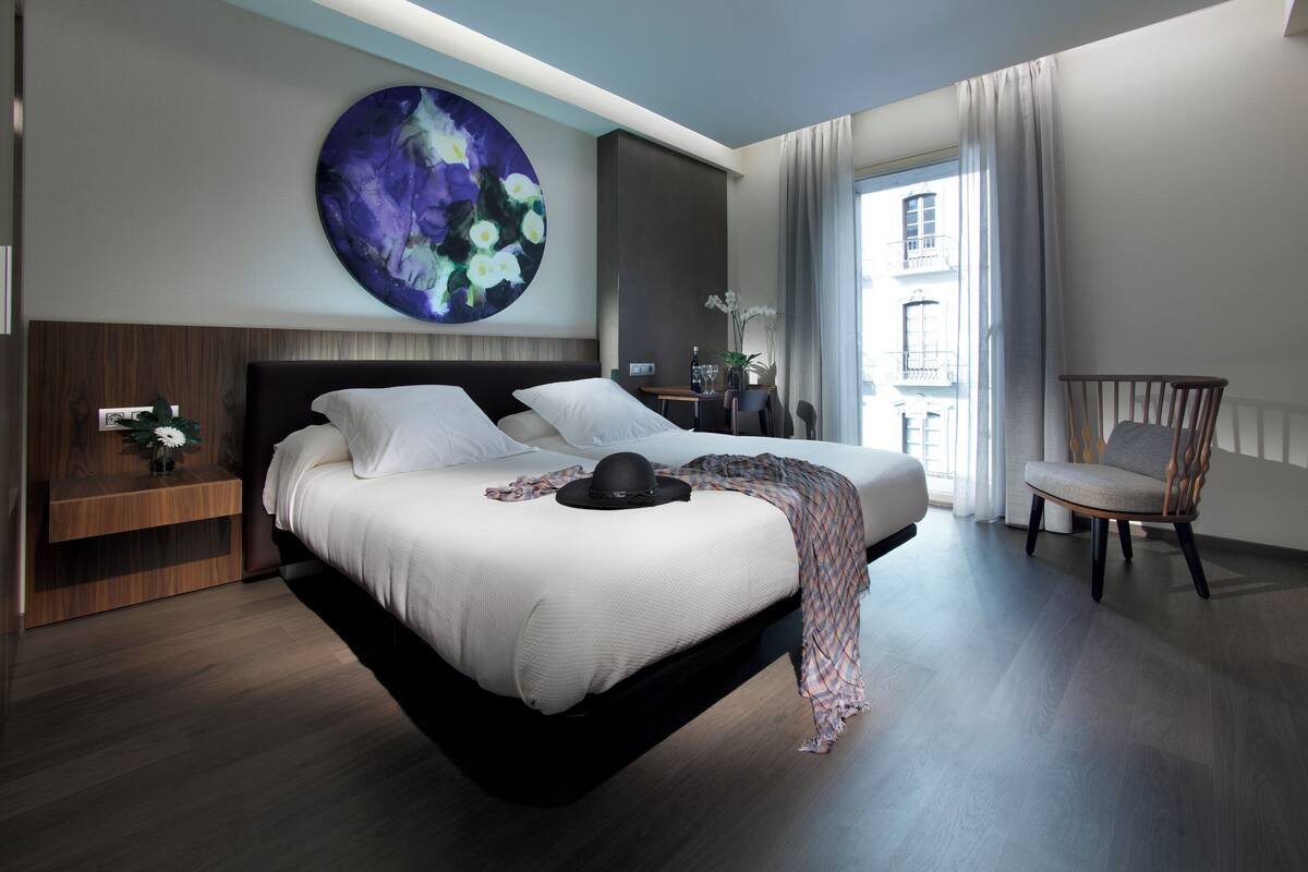 A modern hotel room with a large bed, a nightstand, a chair, and a large abstract painting on the wall.