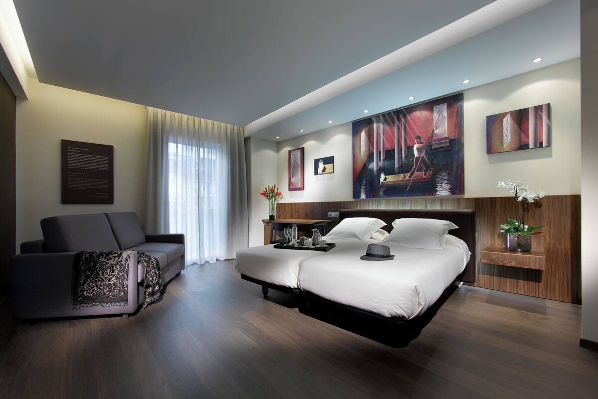 A modern hotel room with a large bed, a sofa, a desk with a tray of items, and artwork on the walls.