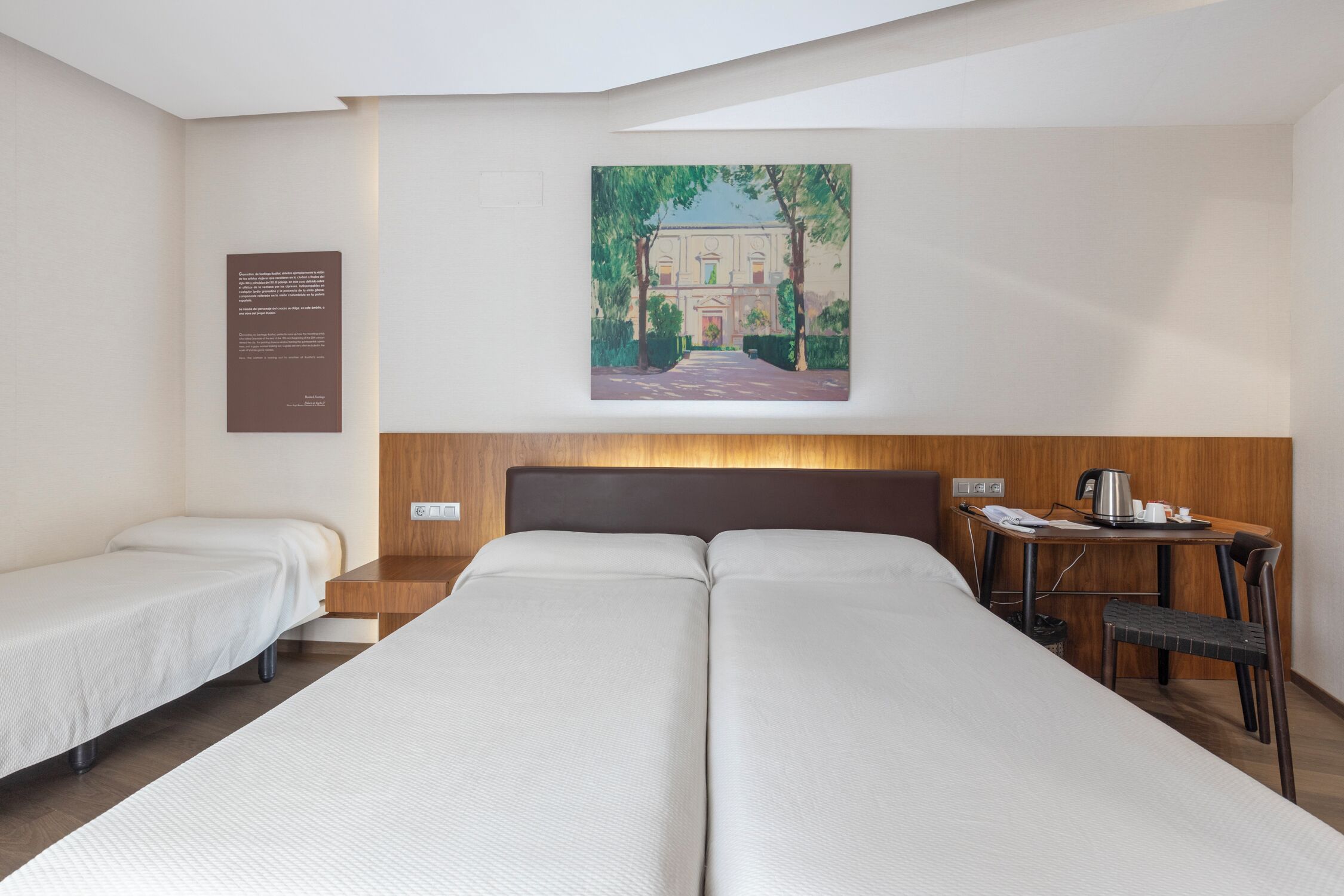 A hotel room with two single beds, a wooden desk, a chair, and a painting on the wall.