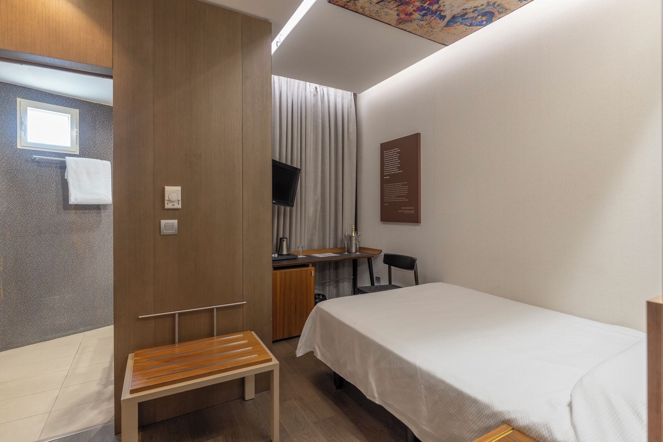 Habitación de hotel compacta con una cama individual, un pequeño escritorio con silla, televisión y ventana con cortinas.