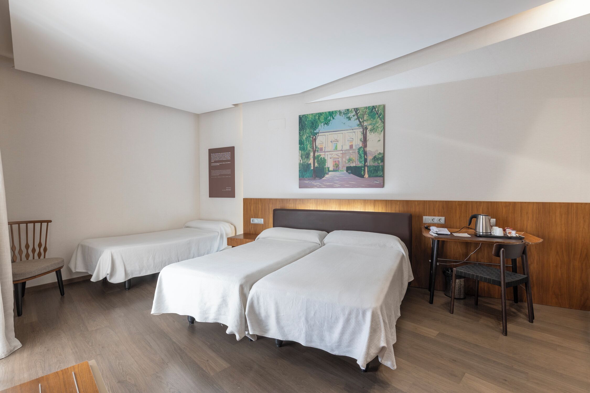 Habitació d'hotel amb dos llits individuals, un panell de fusta al capçal, una taula petita amb una paperera i una cadira.
