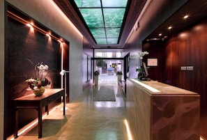 Un moderno vestíbulo de hotel con un mostrador de recepción de mármol, una zona de recepción de madera, un techo de cristal y un largo pasillo con alfombra.
