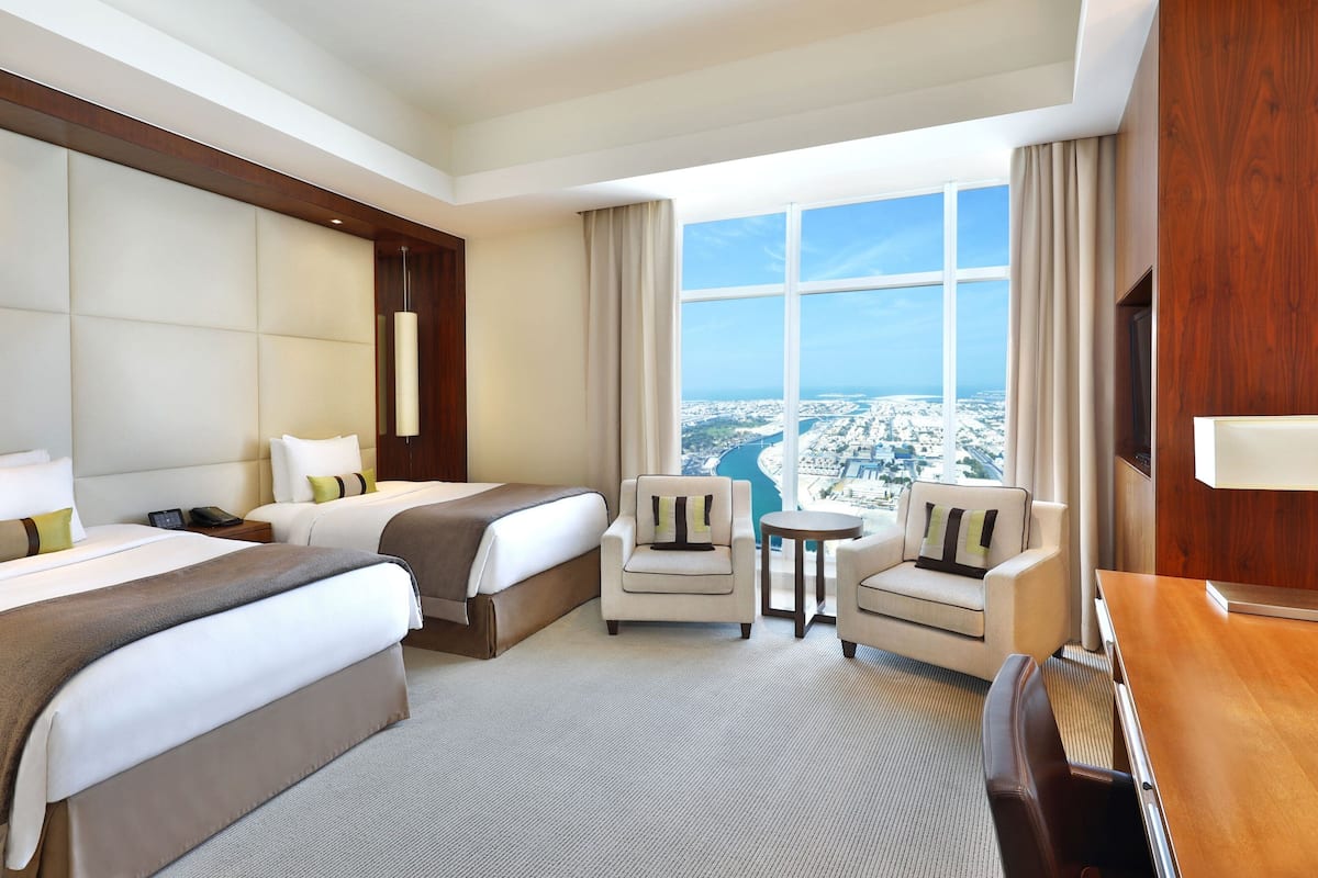A hotel room with two beds, a small table, a chair, and a large window offering a city view.