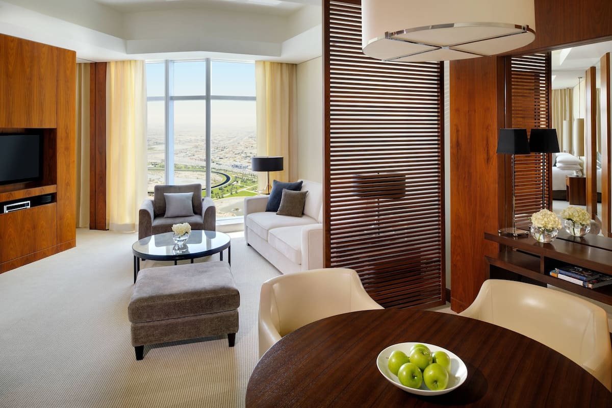 A modern hotel room with a large window offering a city view, a flat-screen TV, a dining area with a table and chairs, and a sofa.
