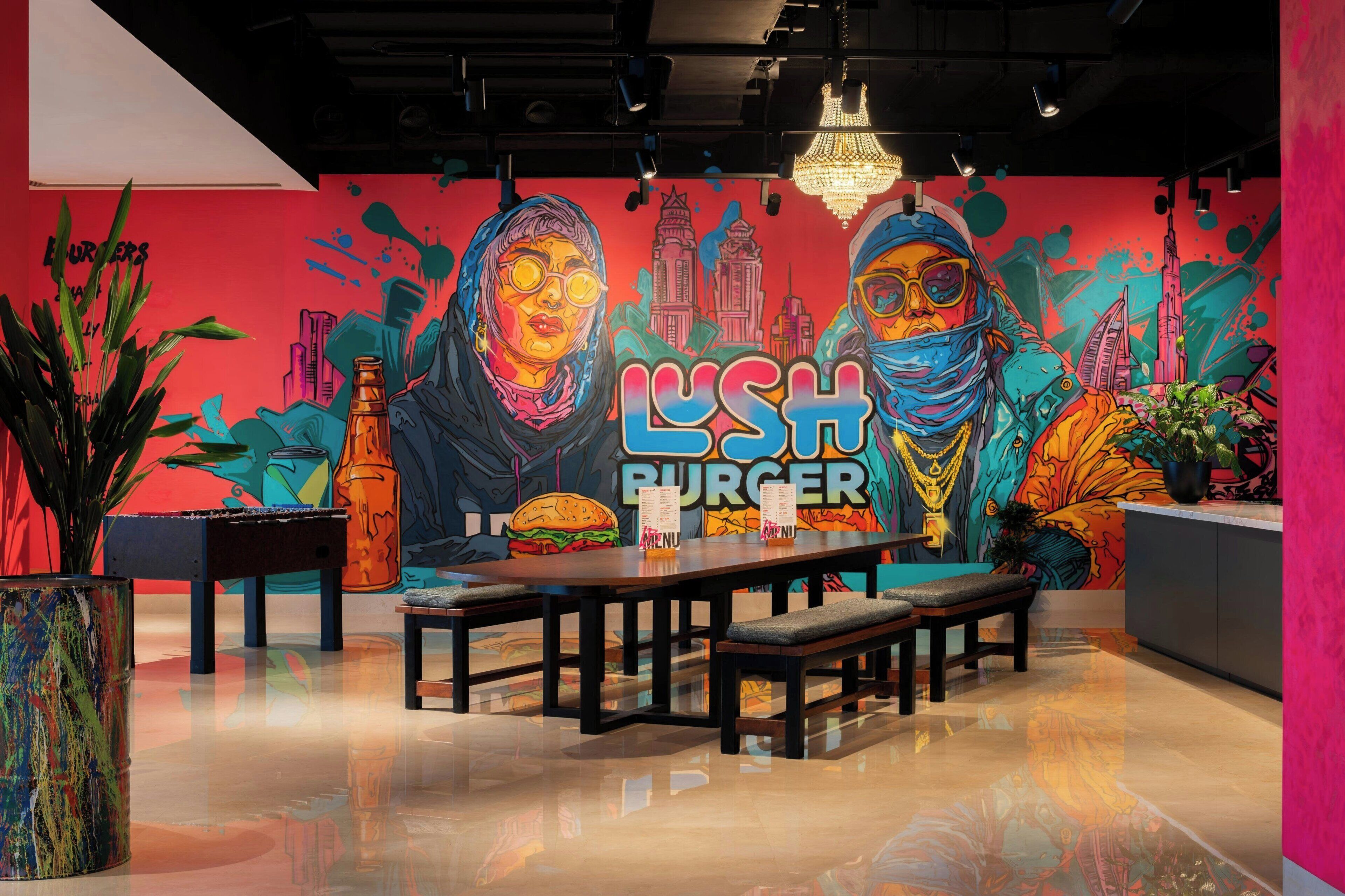 Un interior de restaurante con un mural vibrante, una larga mesa de comedor, bancos y plantas en macetas.