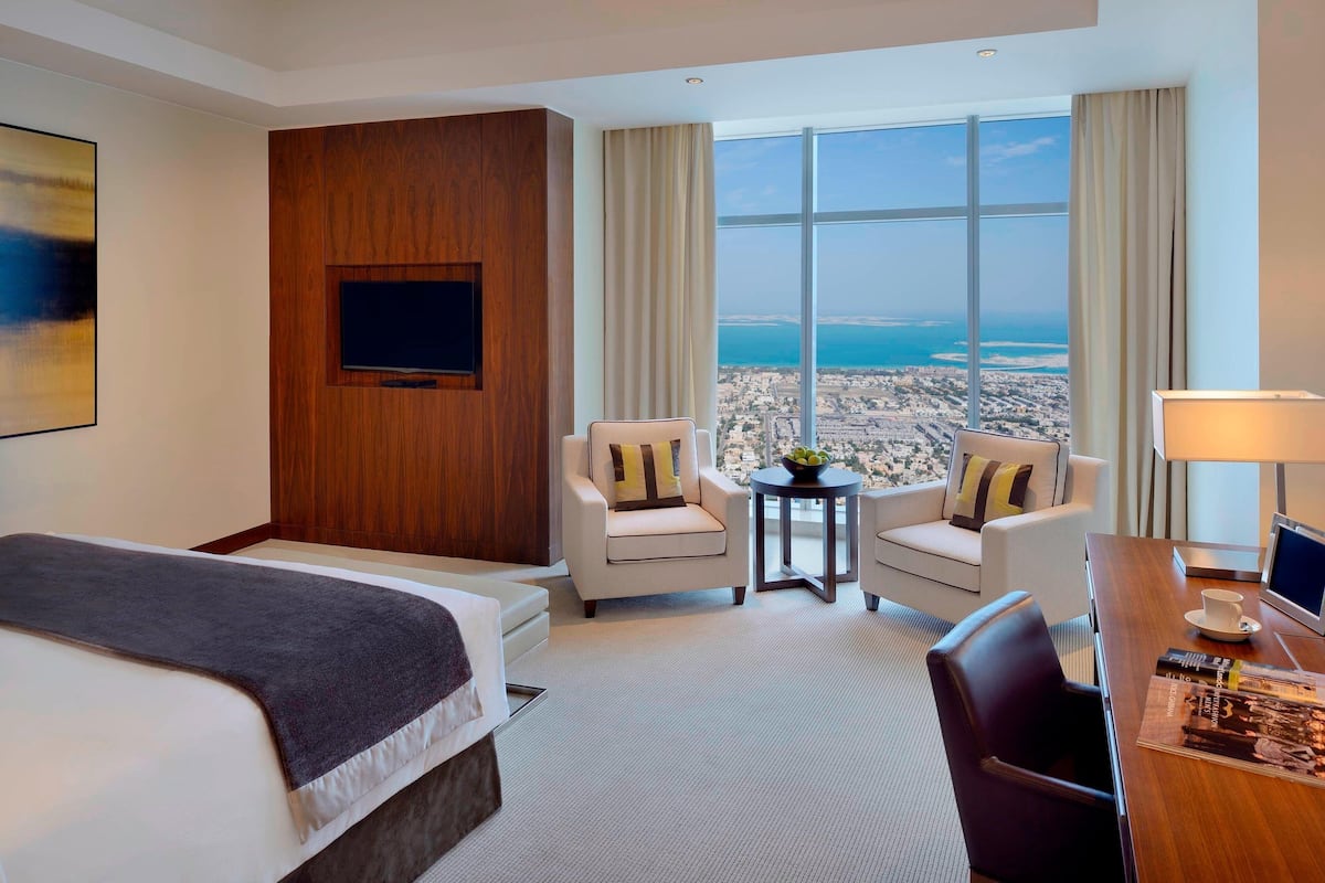 A hotel room with a large bed, two armchairs, a small table, a desk with a lamp, and a TV.