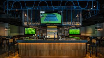 A bar area with a large screen displaying a tennis match, multiple monitors, and a bar counter with stools.