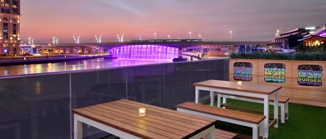 An outdoor seating area with wooden tables and benches, a water feature, and a view of a bridge and buildings in the background.