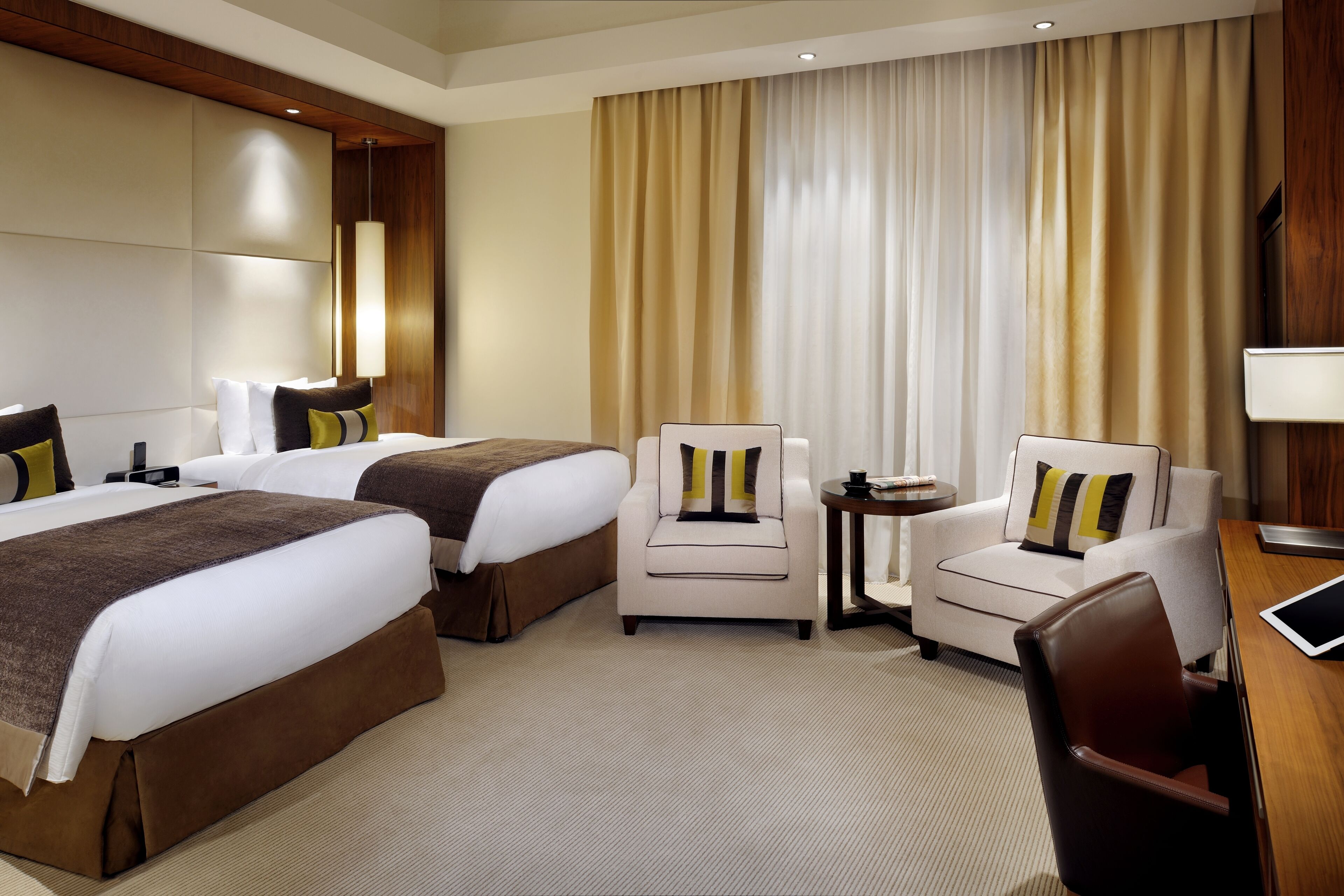 A hotel room with two beds, a sofa, a chair, a small table, and a desk with a lamp.