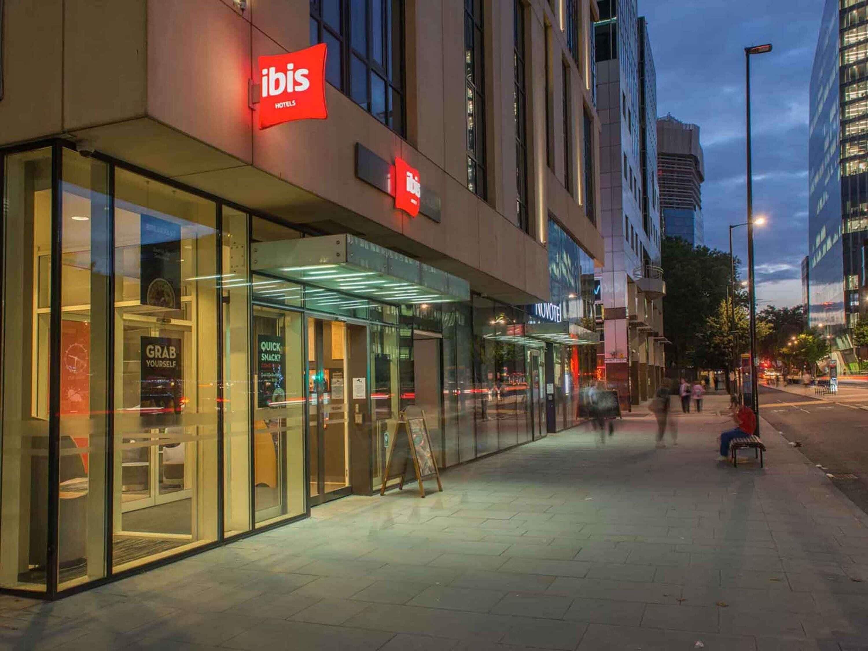 Een modern hotel met glazen gevels, een opvallend 'ibis' bord en een bord met de tekst 'haal iets voor jezelf', wat wijst op een self-service snackgedeelte.