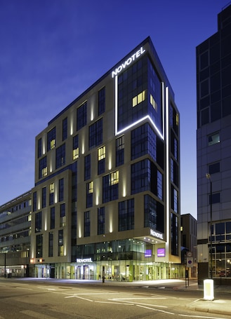 A modern Novotel hotel with a prominent 'Novotel' sign on the facade, large windows, and a well-lit entrance area.