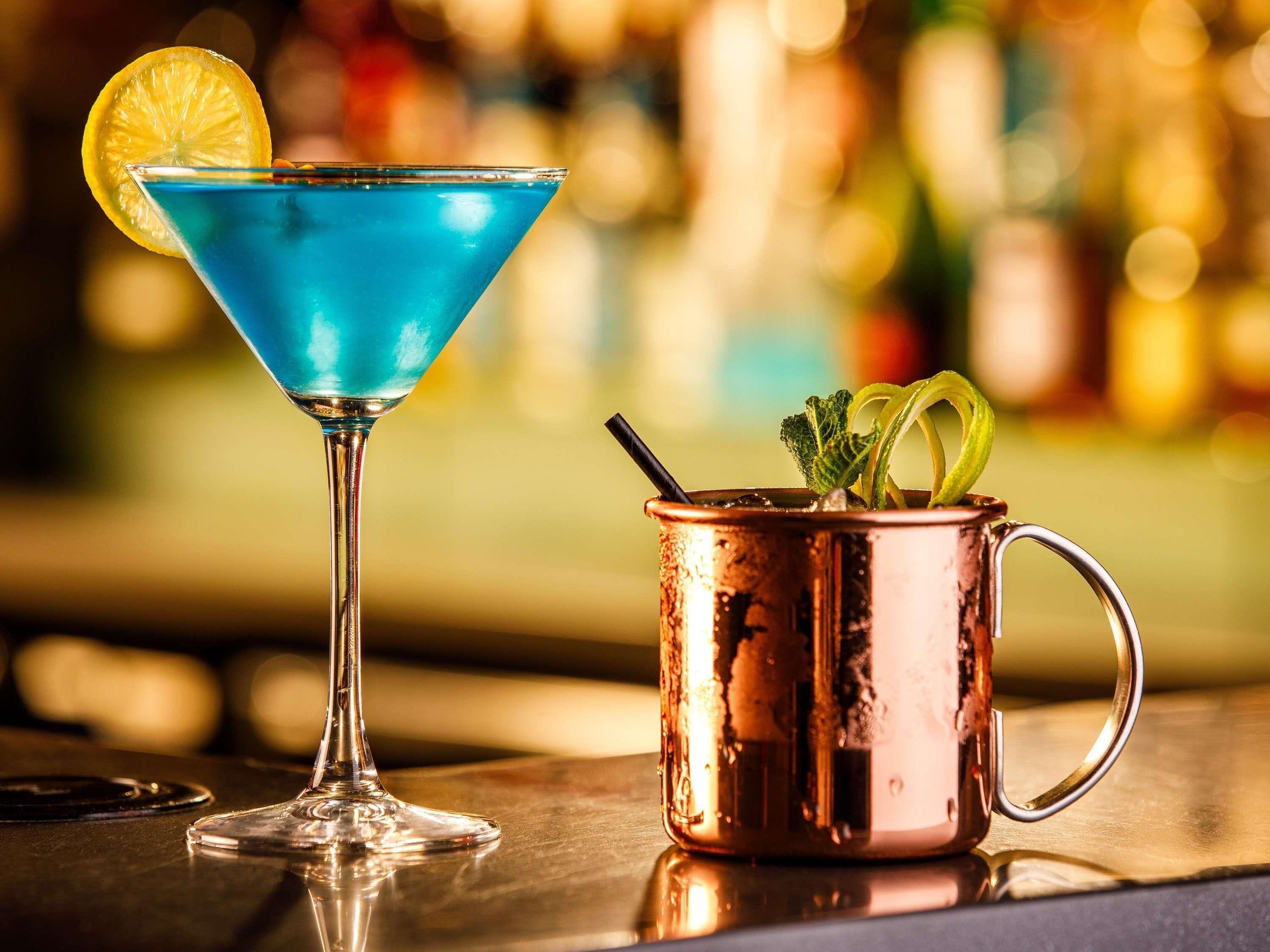 Two drinks, one in a martini glass and the other in a copper mug, on a bar counter with a blurred background of bottles.