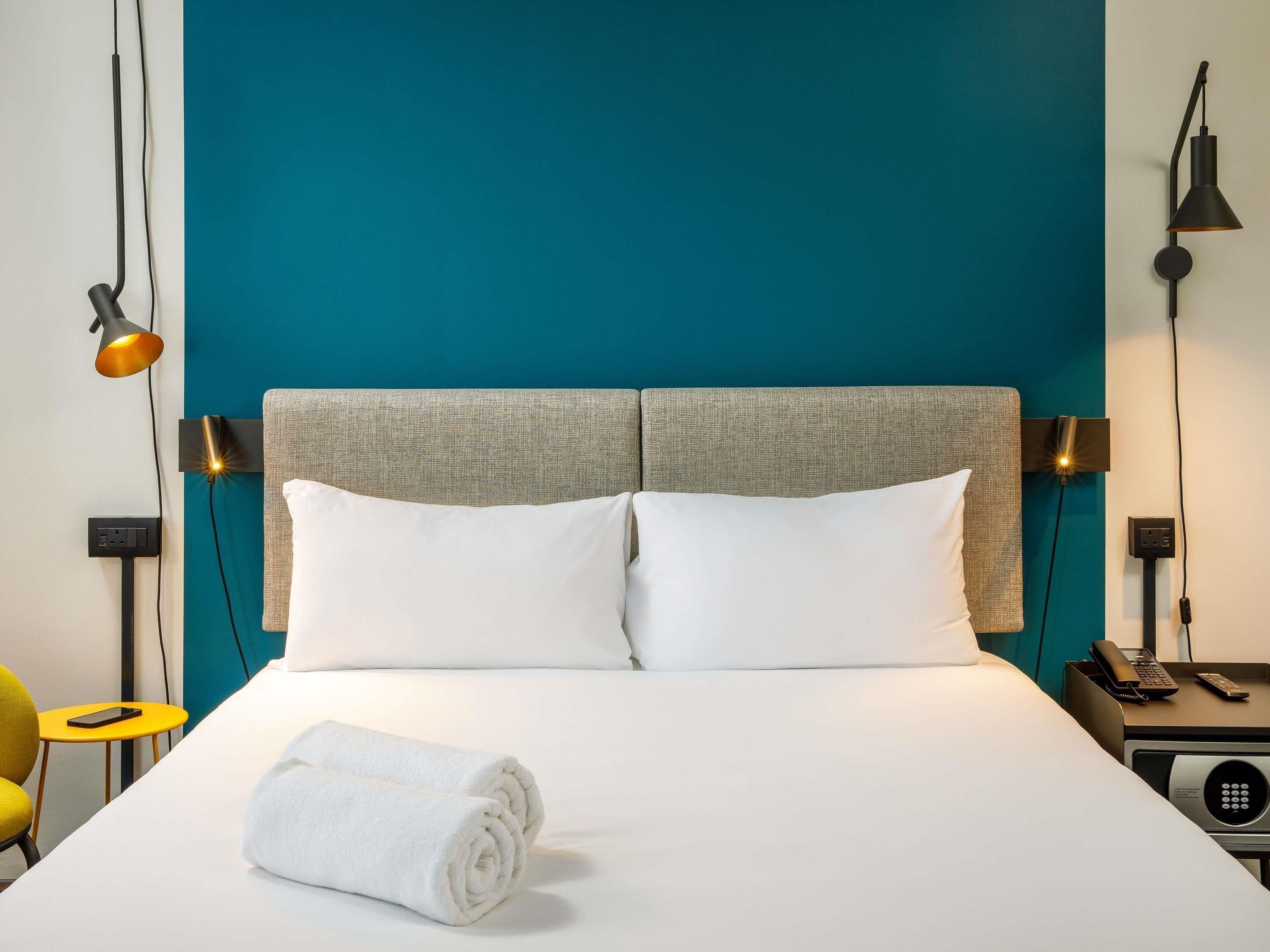 A hotel room with a teal accent wall, a bed with white linens, two bedside lamps, a bedside table with a phone and remote, and a rolled white towel on the bed.