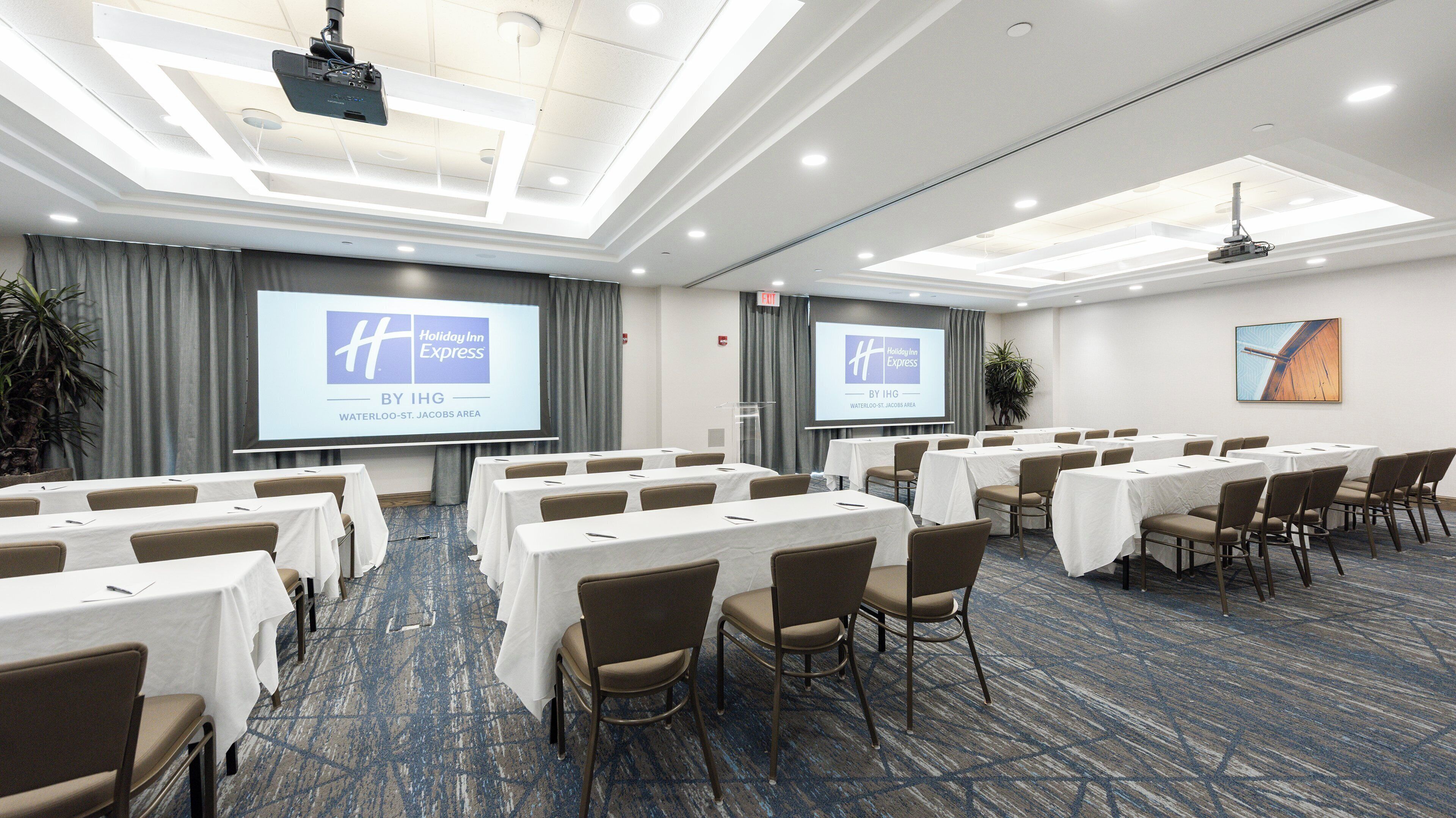 Un salón de conferencias con filas de mesas y sillas, una pantalla de proyector mostrando una presentación y una vista clara del logotipo del hotel.