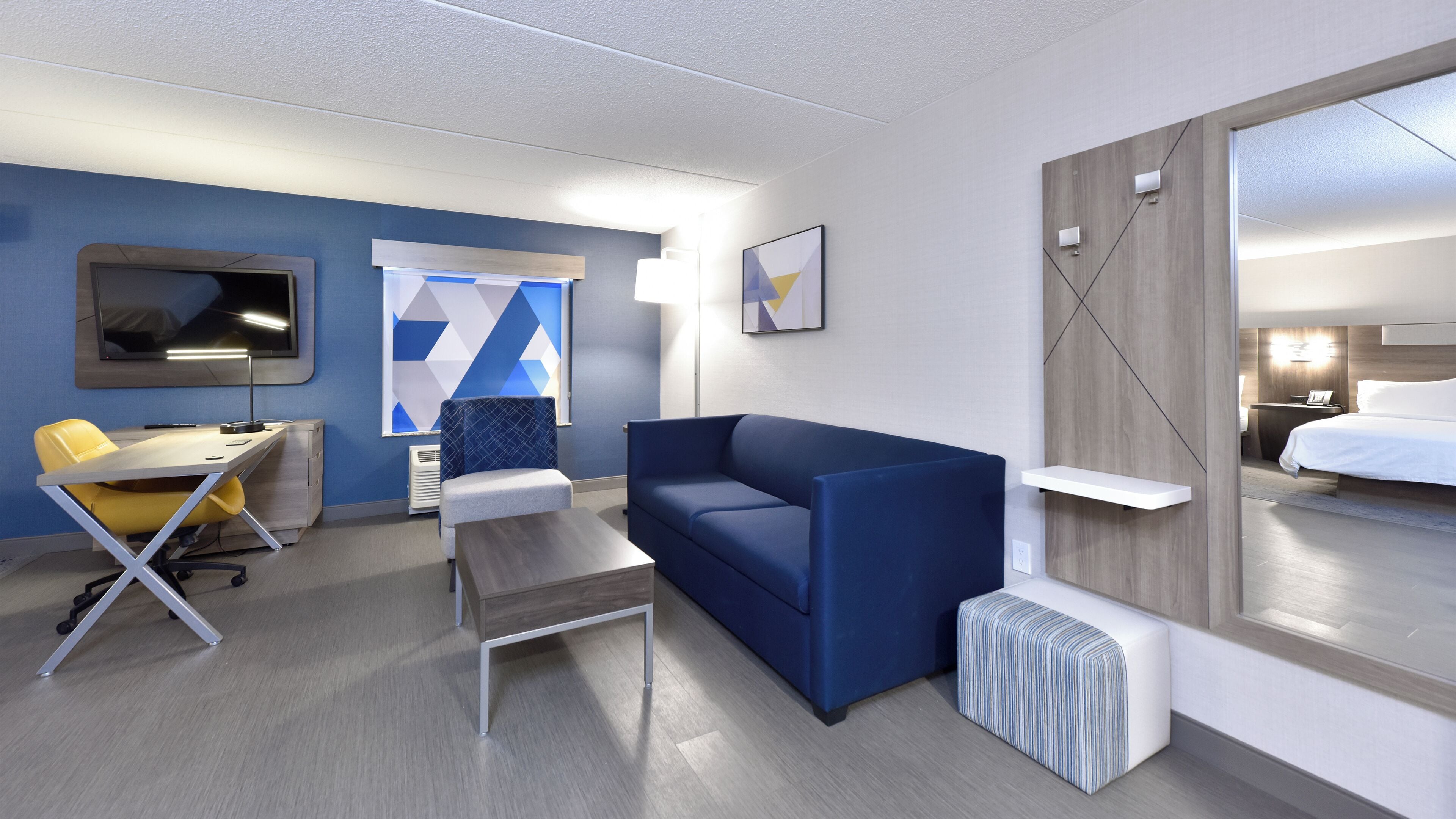 A modern hotel room with a blue sofa, a yellow chair, a desk, and a bed.
