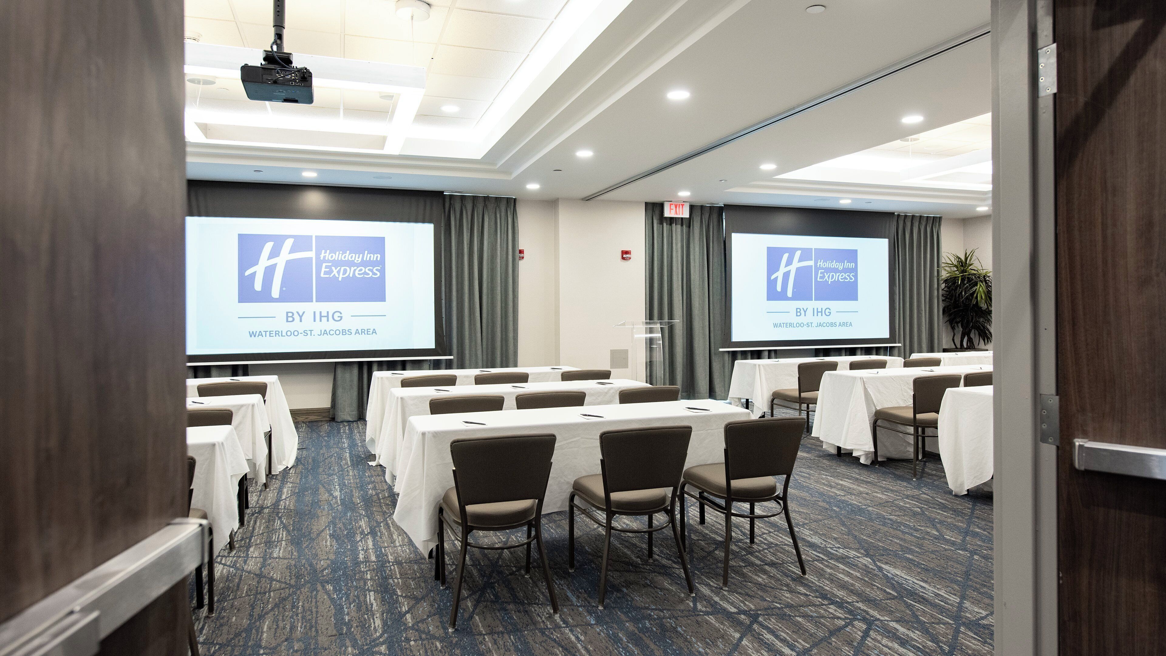 Ein Konferenzraum mit Tischen, Stühlen und einer Projektionsleinwand, die ein Holiday Inn Express Logo zeigt.