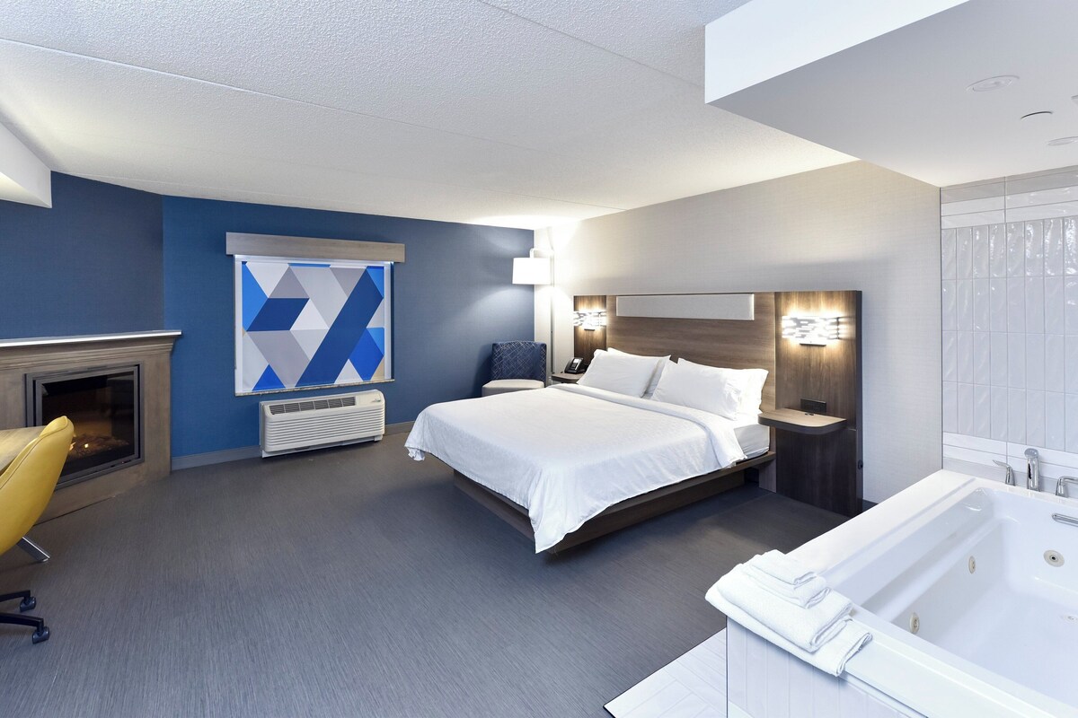 A modern hotel room with a large bed, a fireplace, a sitting area with a yellow chair, a desk, and a bathroom with a bathtub.