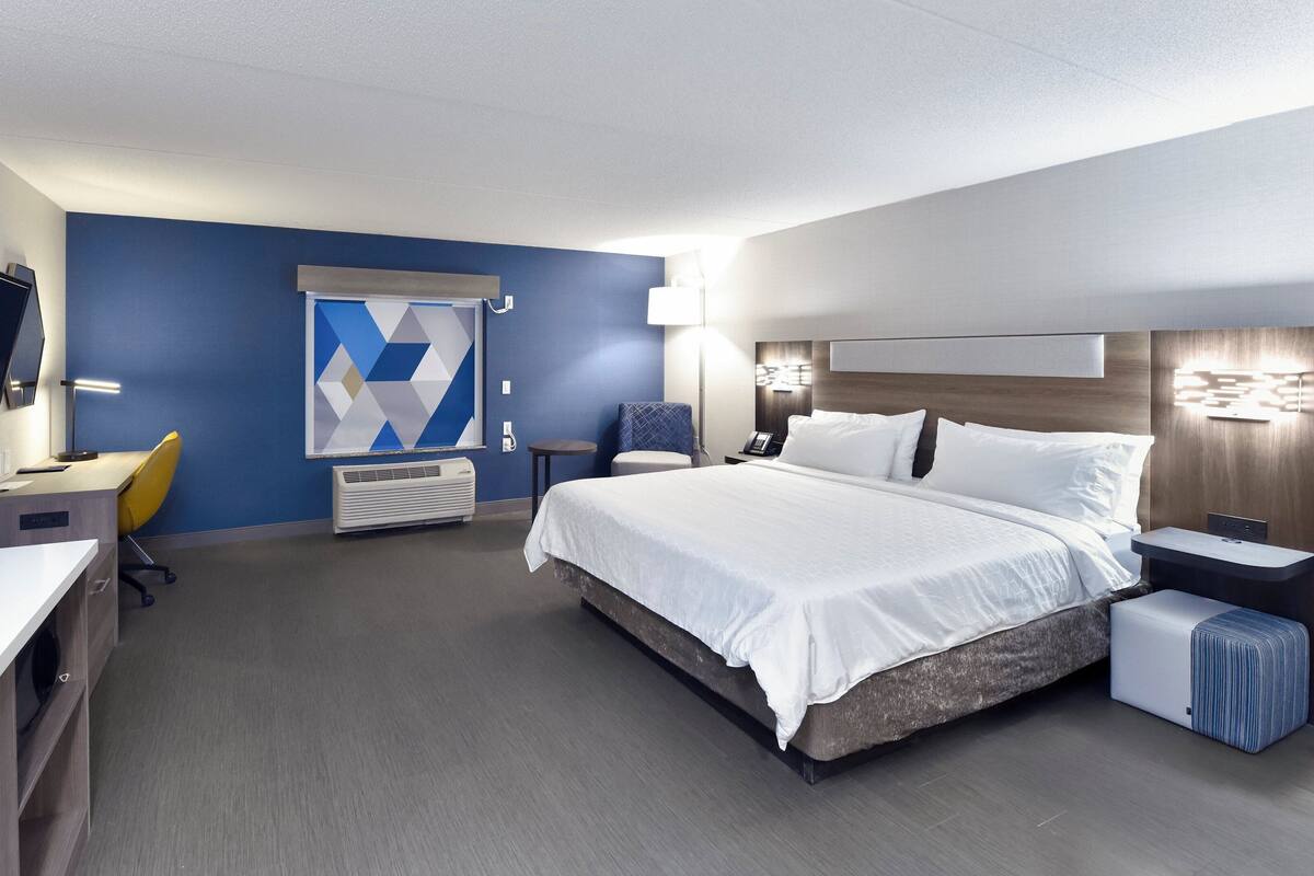 A modern hotel room with a large bed, a desk, a chair, a TV, and a decorative wall piece.