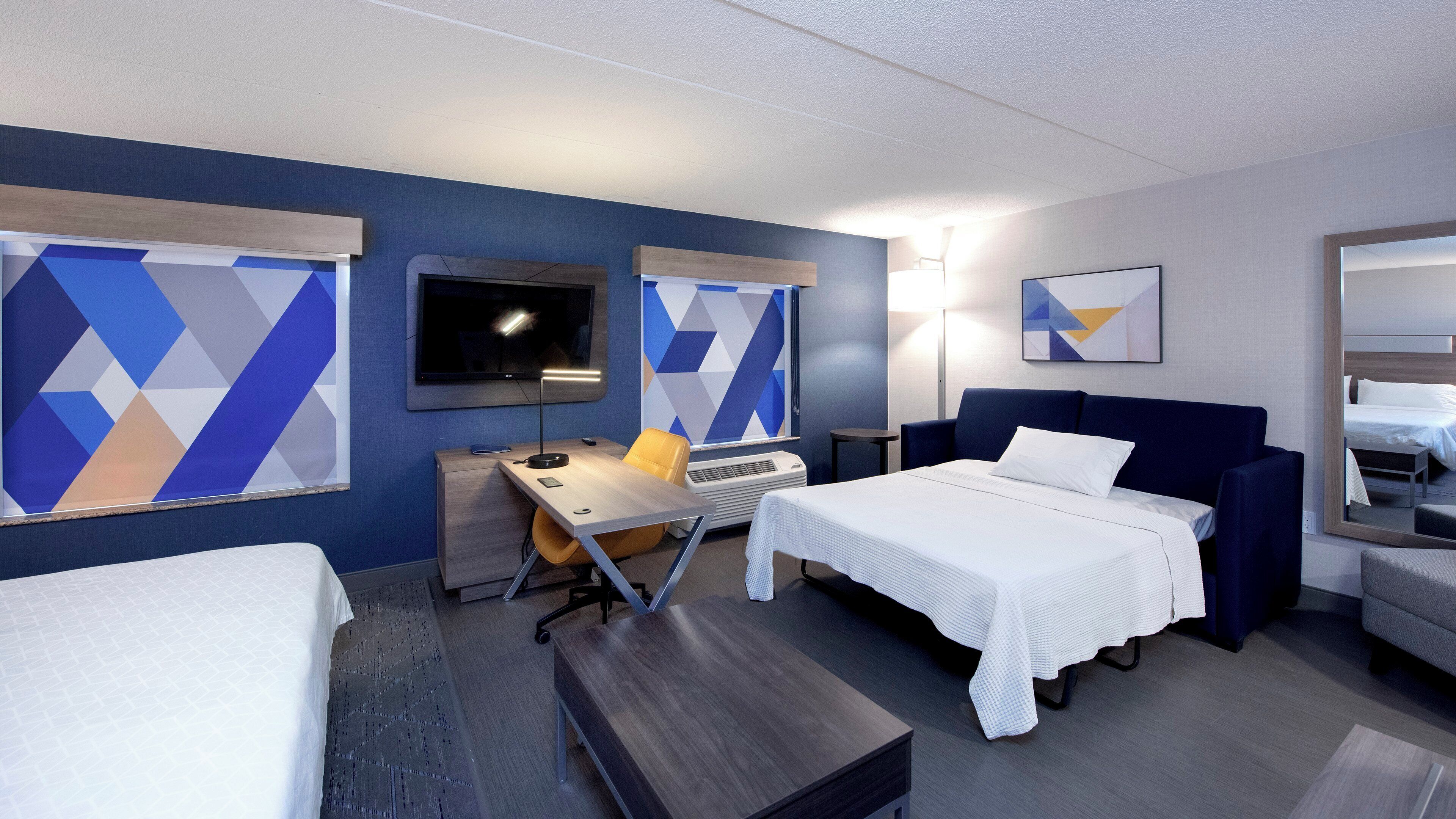 Une chambre d’hôtel comprenant un lit, un bureau, une chaise, un téléviseur et une grande fenêtre ornée de motifs géométriques.