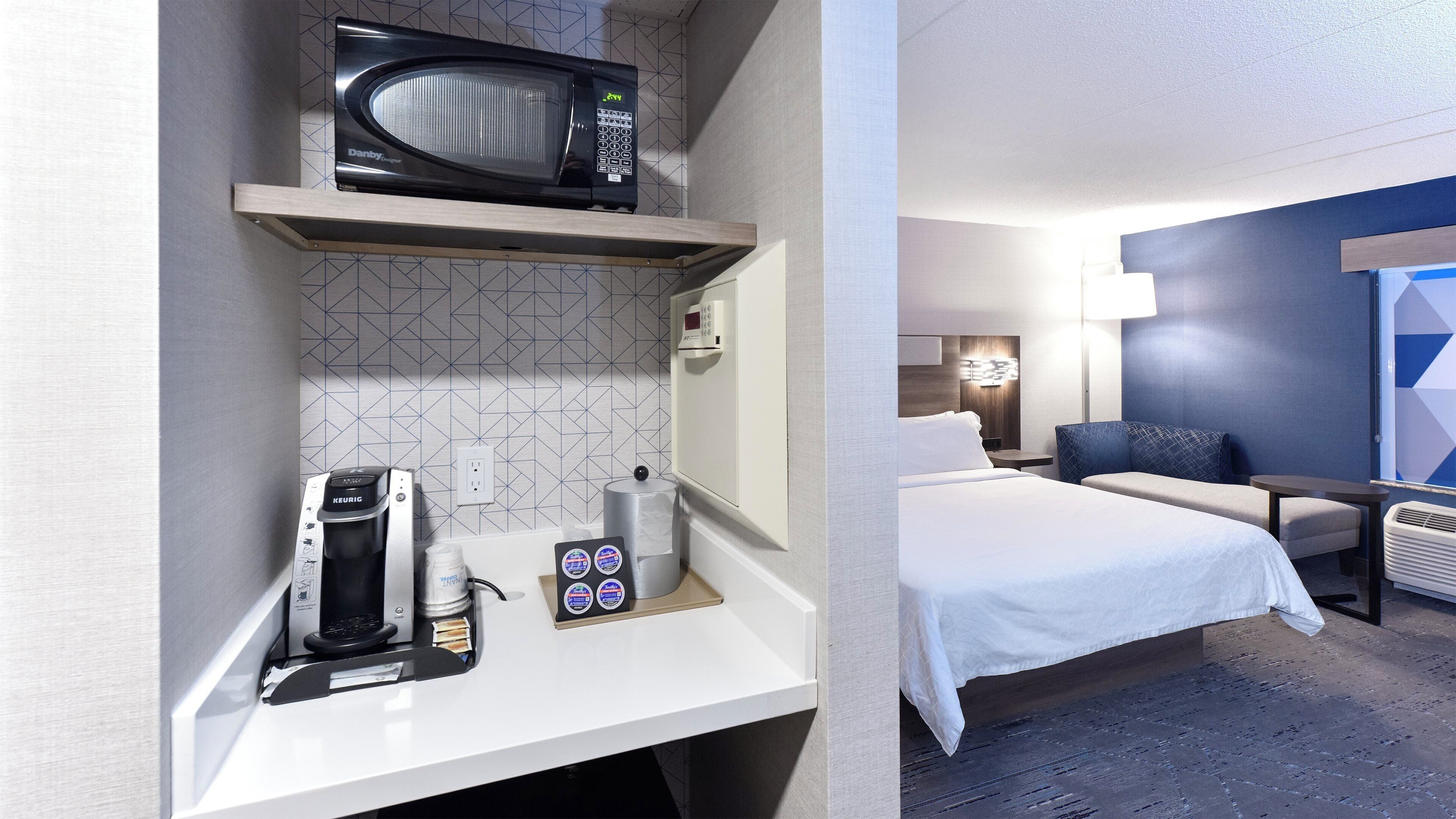 A hotel room with a bed, a desk with a coffee maker, a microwave, and a small table with a chair.