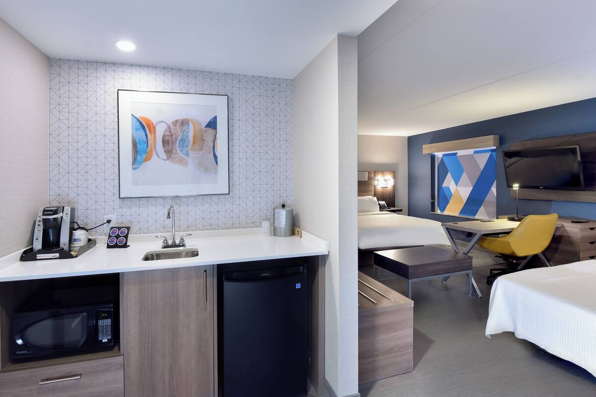 A modern hotel room with a kitchenette, a bed, a desk, and a TV.