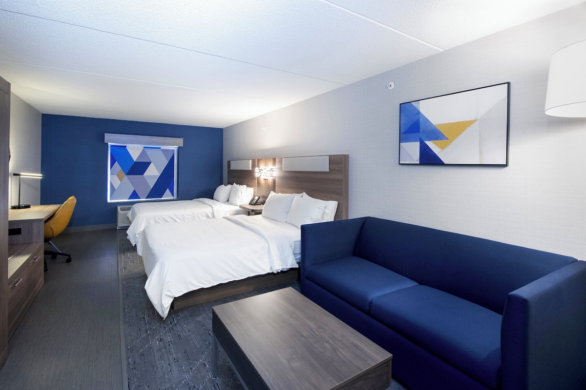 A modern hotel room with a large bed, a blue sofa, a desk with a chair, and a desk lamp.
