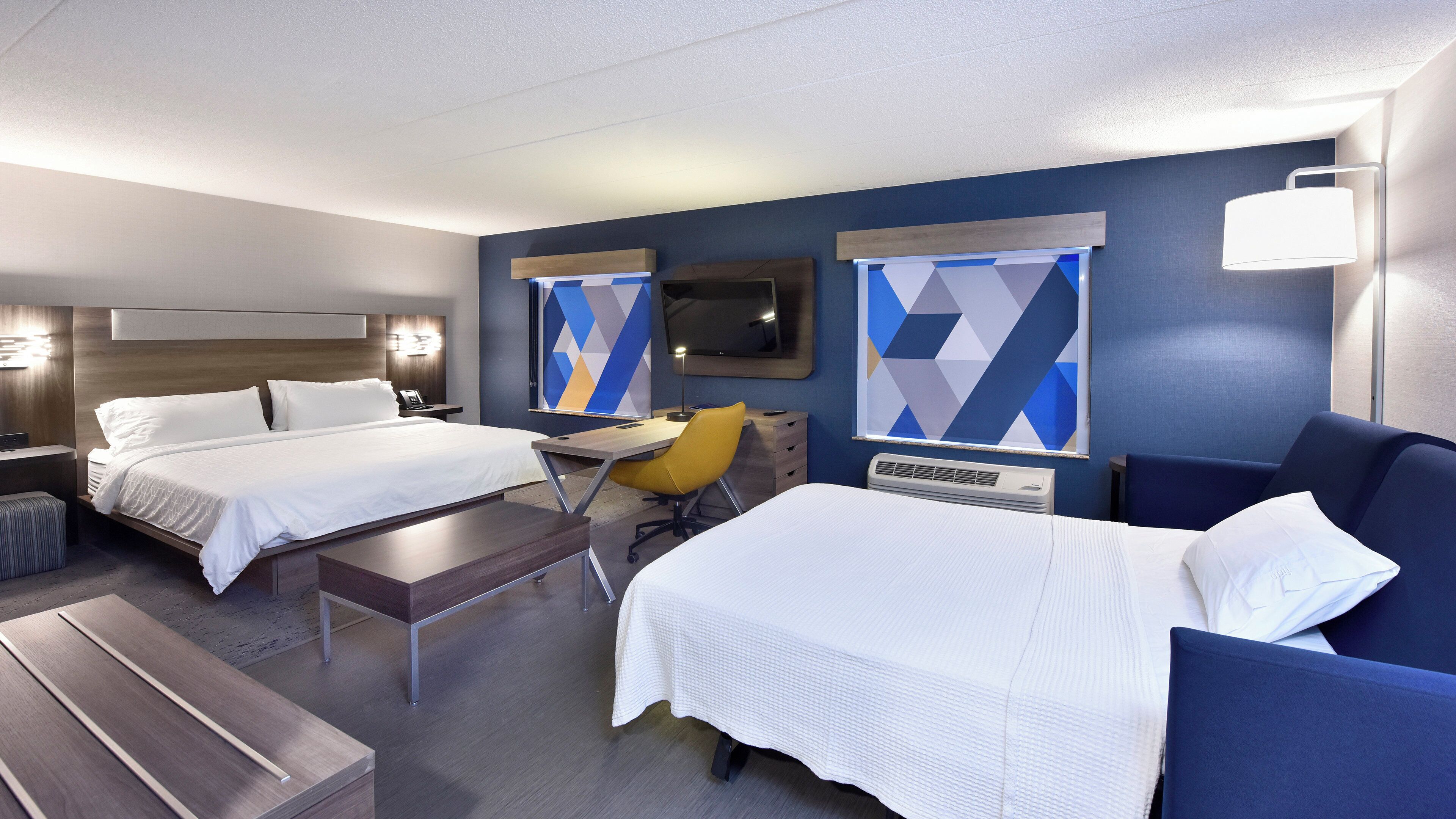 Quarto de hotel com duas camas, uma mesa, uma cadeira, uma televisão e uma janela grande com cortinas geométricas.