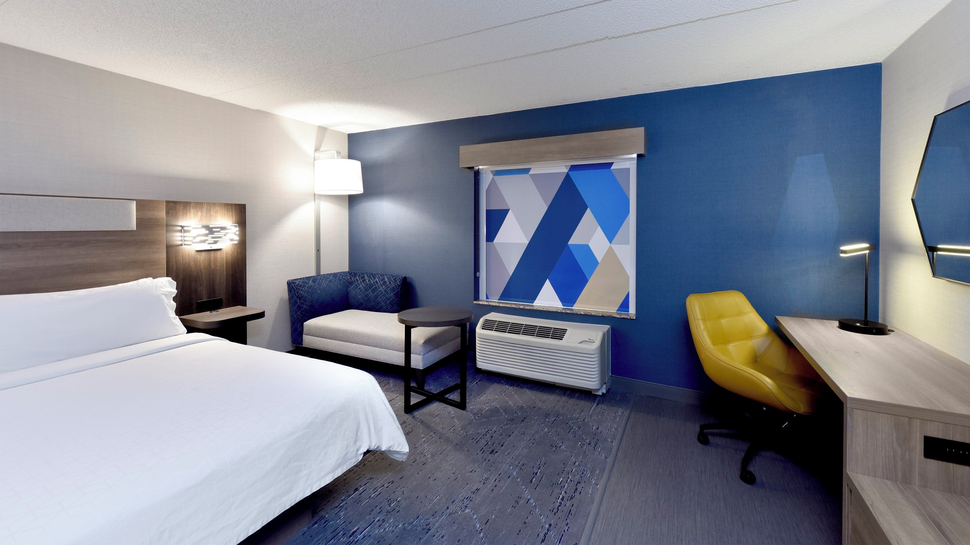 A hotel room with a bed, a desk, a chair, a heater, and a large window with a geometric view.