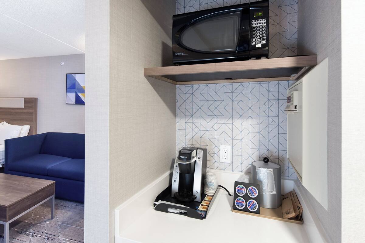 A hotel room with a bed, a blue sofa, a wooden coffee table, a microwave, a coffee machine, and a small refrigerator.