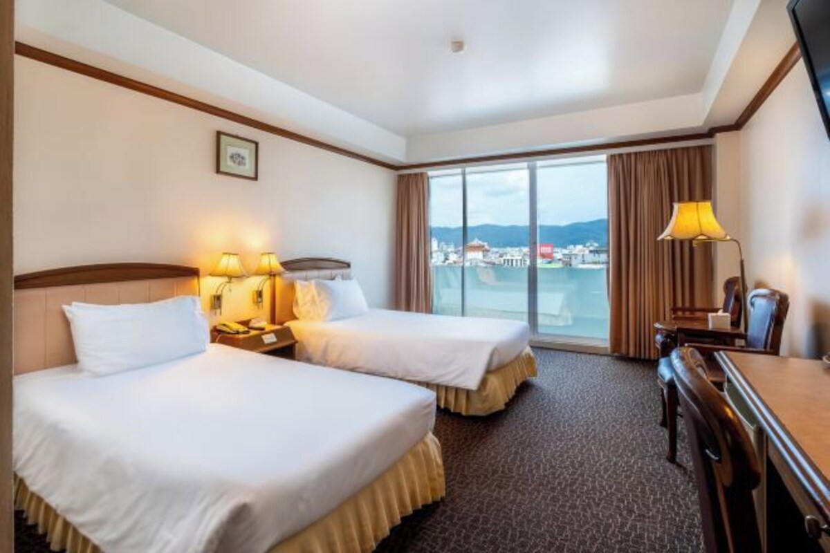 A hotel room with two beds, a desk, and a large window offering a view of a cityscape.