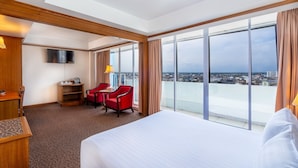 A hotel room with a large bed, a desk with a chair, a TV, and a city view through large windows. - V.L. HatYai Hotel (Hat Yai)