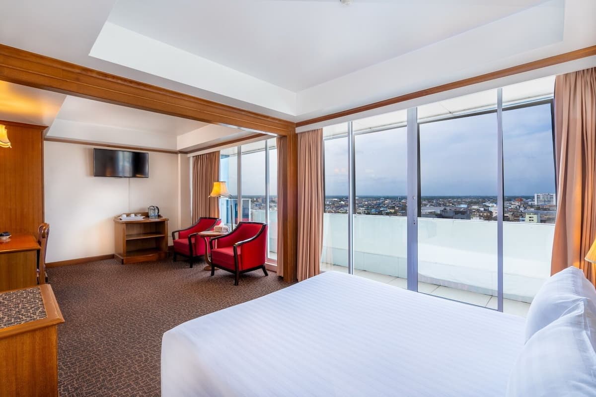 Habitación de hotel con una cama grande, un escritorio con silla, televisión y vistas a la ciudad a través de amplias ventanas.