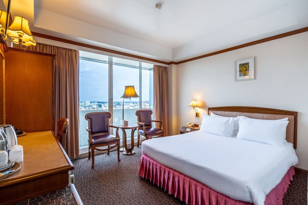 Habitación de hotel con una cama grande, dos sillones, una mesita, un escritorio con lámpara y vistas a la ciudad a través de amplios ventanales.