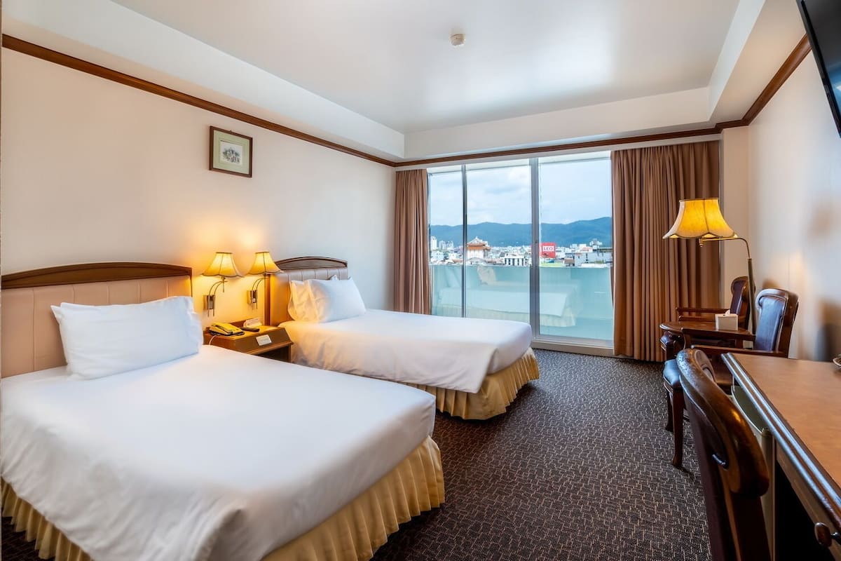 Habitación de hotel con dos camas, un escritorio, una silla, una lámpara y un amplio ventanal con vistas a la ciudad.