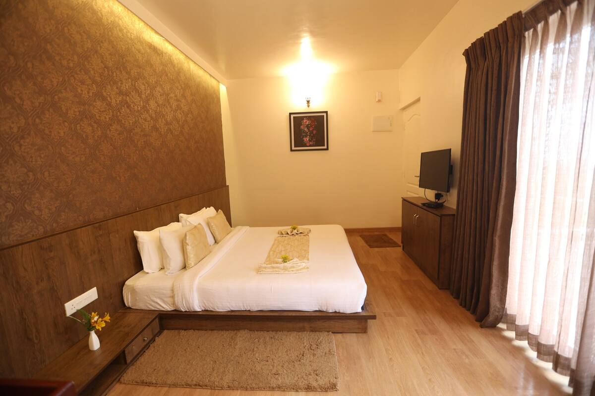 A hotel room with a large bed, a flat-screen TV, a wooden headboard, a small desk with a vase of flowers, and a large window with curtains.