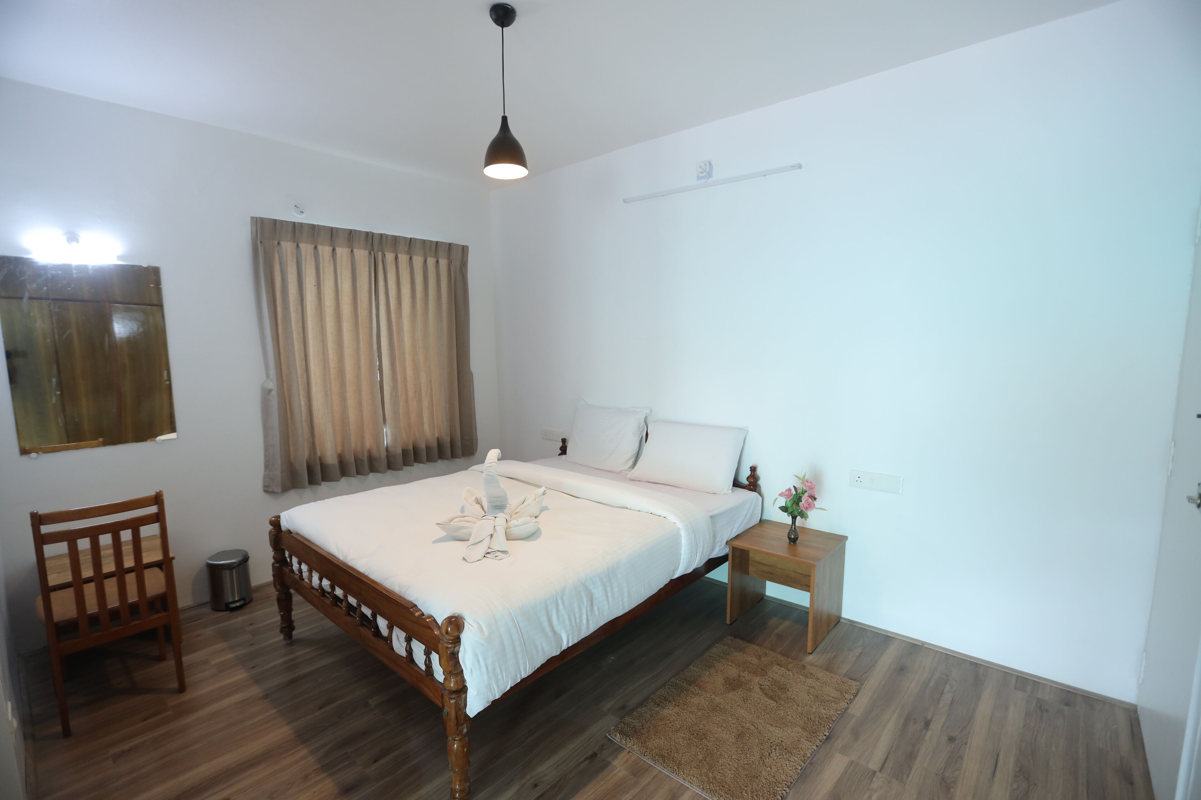 A bedroom with a wooden bed, a bedside table, a chair, a small trash bin, and a window with curtains.