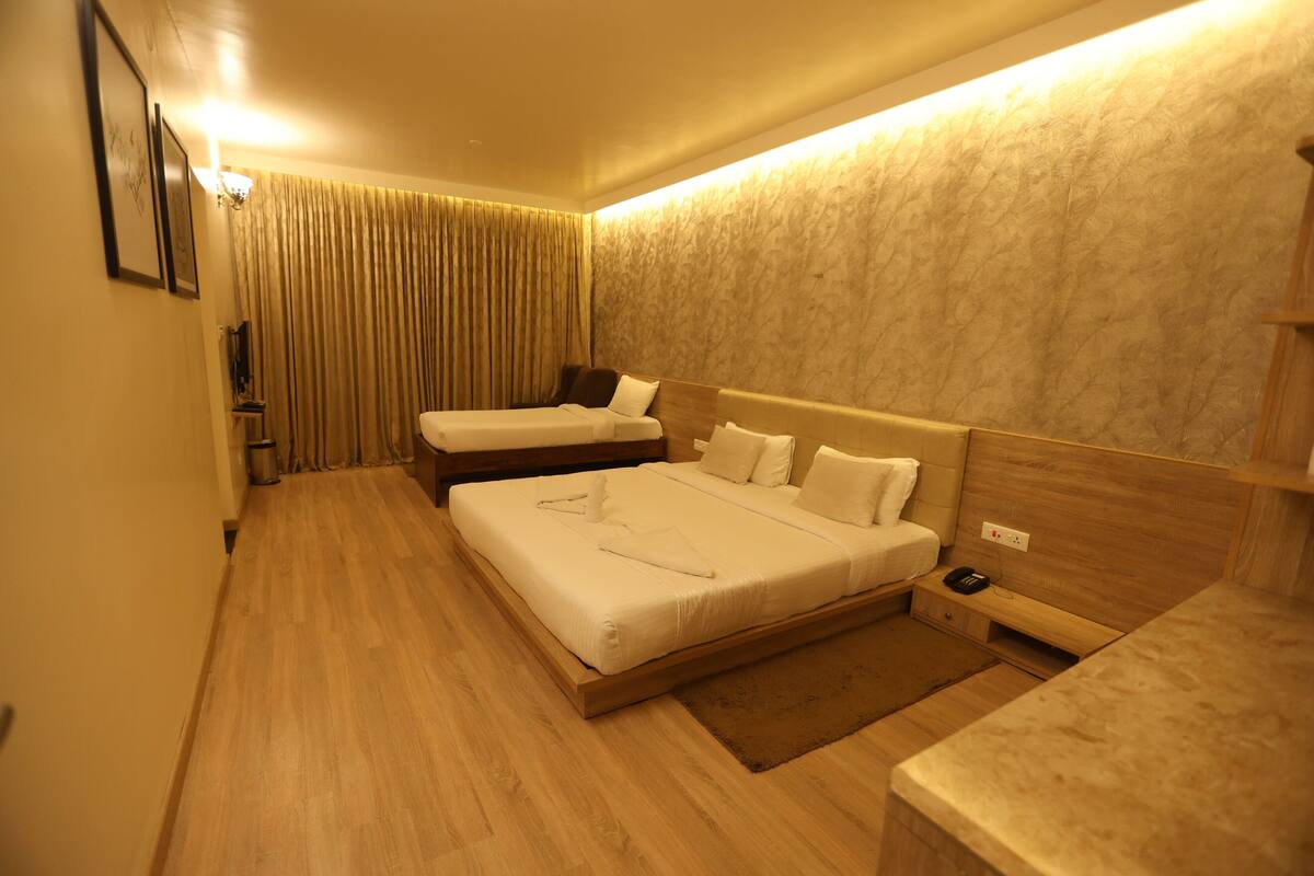 A hotel room with a large bed, a smaller bed, a bedside table, a nightstand, a lamp, and a curtain.
