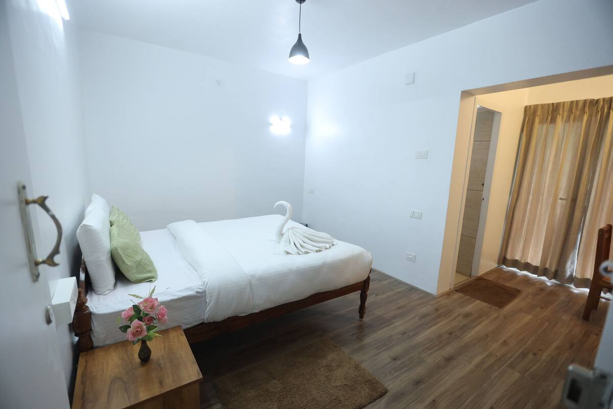 A bedroom with a wooden bed, a bedside table with a vase of flowers, a brown rug, and a door leading to another room.